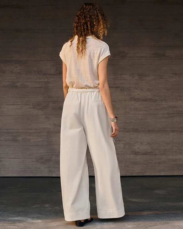A woman viewed from behind wearing a light beige short-sleeve top and off-white wide-leg trousers, standing against a dark gray wood-paneled wall.
