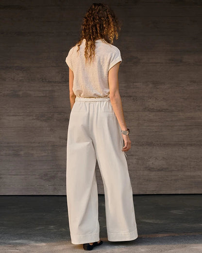 A woman viewed from behind wearing a light beige short-sleeve top and off-white wide-leg trousers, standing against a dark gray wood-paneled wall.