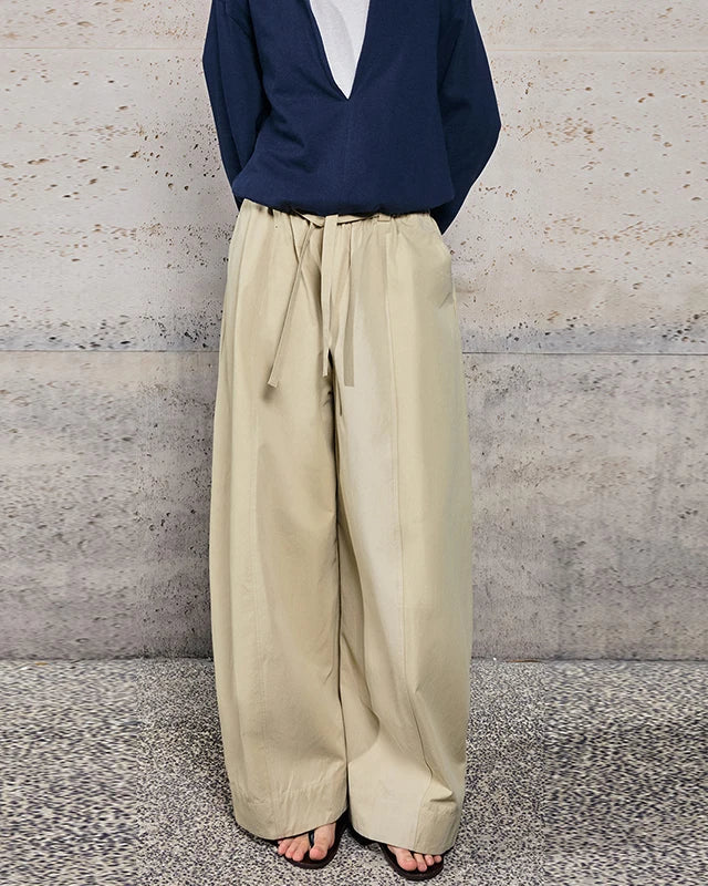 A person wearing wide-leg beige drawstring pants with a navy blue top, standing against a textured concrete wall.