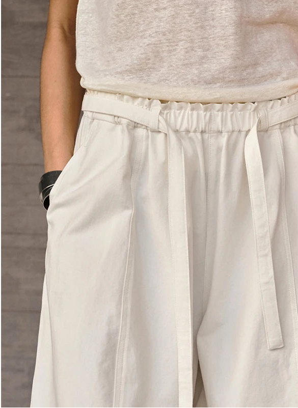 Close-up of a person wearing off-white wide-leg trousers with a matching drawstring waistband, paired with a light beige top. The person is standing against a textured gray wall.