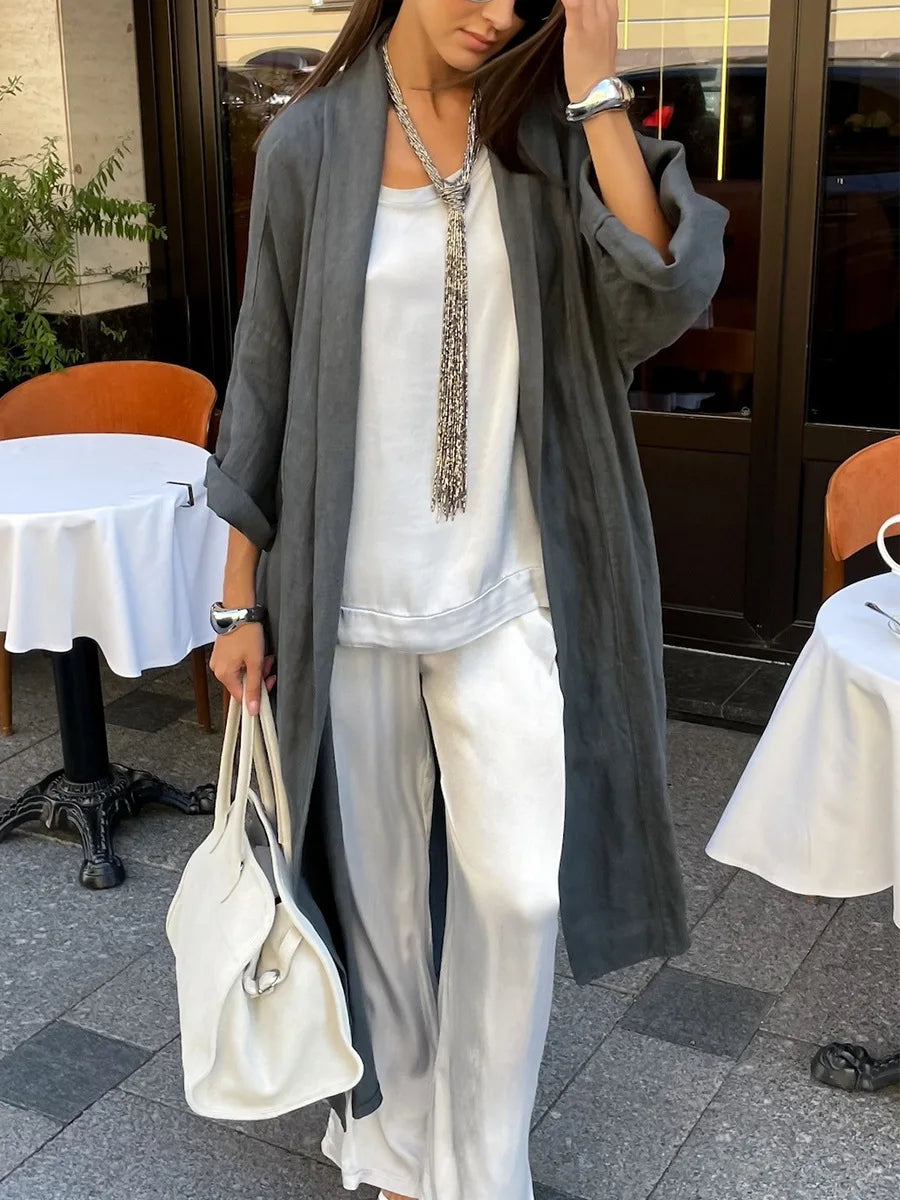 A woman wearing a gray open-front cardigan over a white top and white wide-leg pants, accessorized with a long silver beaded tassel necklace and carrying a white handbag, standing on a patio.