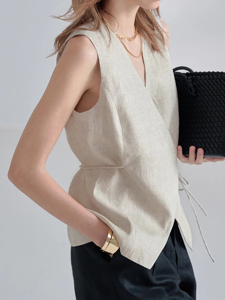 A woman wearing a sleeveless beige linen wrap vest with a deep V-neck and side tie detail, paired with black trousers and holding a black woven clutch bag.