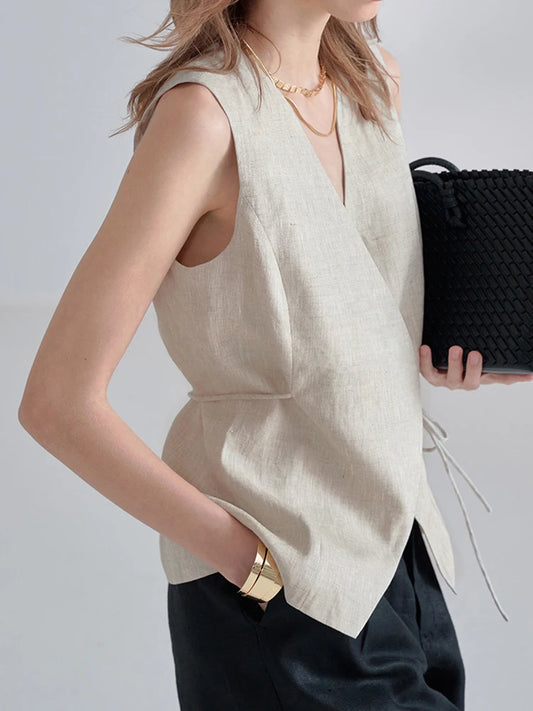 A woman wearing a sleeveless beige linen wrap vest with a deep V-neck and side tie detail, paired with black trousers and holding a black woven clutch bag.