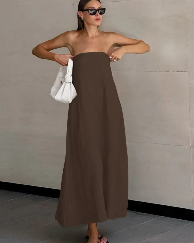 Woman in a brown strapless dress holding a white bag against a plain wall.