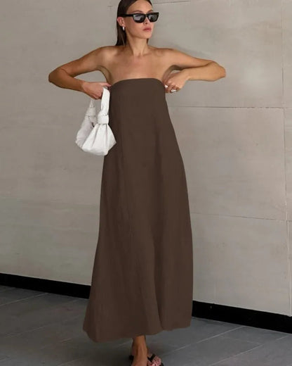 Woman in a brown strapless dress holding a white bag against a plain wall.
