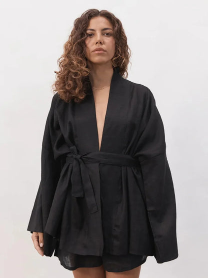 A woman with curly brown hair wearing a black wrap-style robe with a tie waist and long wide sleeves, photographed against a plain light gray studio background.