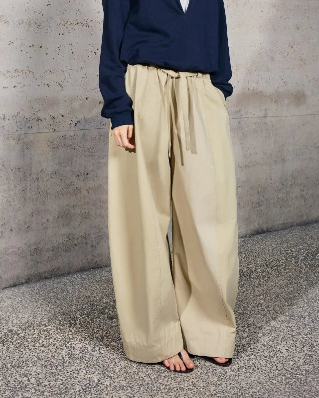 A person wearing loose, wide-leg beige drawstring pants and a navy long-sleeve top, standing barefoot against a textured concrete wall and speckled floor.