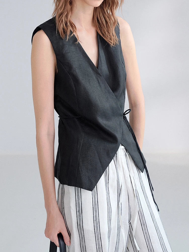 A model wearing a sleeveless black wrap-style vest with a side tie detail, paired with white and black vertically striped trousers, against a light gray studio background.