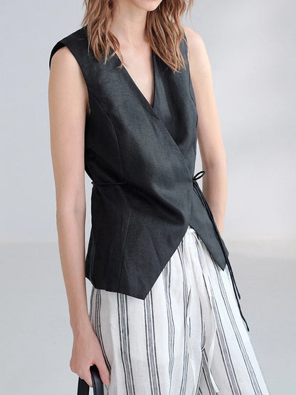 A model wearing a sleeveless black wrap-style vest with a side tie detail, paired with white and black vertically striped trousers, against a light gray studio background.