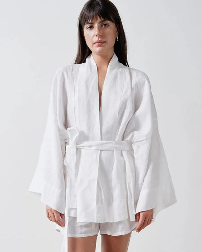A woman modeling a white long-sleeve kimono-style robe with a self-tie belt and matching white shorts against a plain light gray studio background.