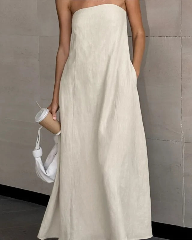 A woman wearing a strapless, floor-length off-white linen maxi dress with side pockets, holding a white shoulder bag and a paper coffee cup against a gray tiled wall.