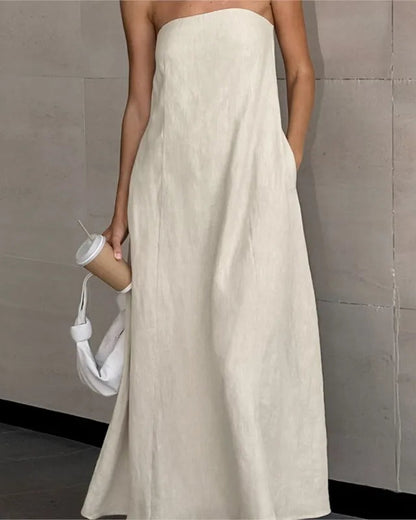 A woman wearing a strapless, floor-length off-white linen maxi dress with side pockets, holding a white shoulder bag and a paper coffee cup against a gray tiled wall.