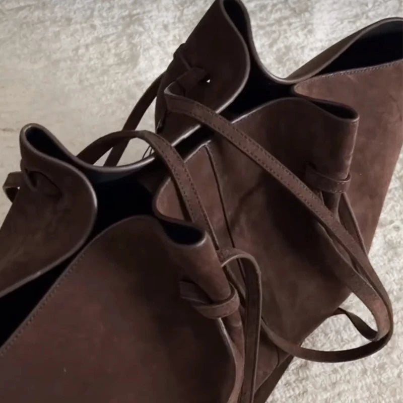 A close-up of a dark brown suede or faux-suede bucket bag with matching shoulder straps, photographed on a light textured surface.