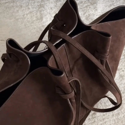 A close-up of a dark brown suede or faux-suede bucket bag with matching shoulder straps, photographed on a light textured surface.