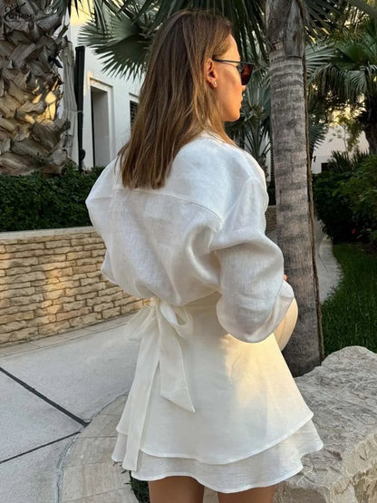 Woman in a white dress standing outdoors with palm trees and a stone wall in the background