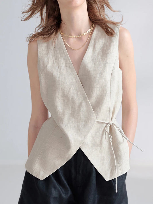 A woman wearing a sleeveless wrap-style vest in a light beige linen-like fabric with a side tie, paired with dark pants and layered gold necklaces, against a plain light gray background.